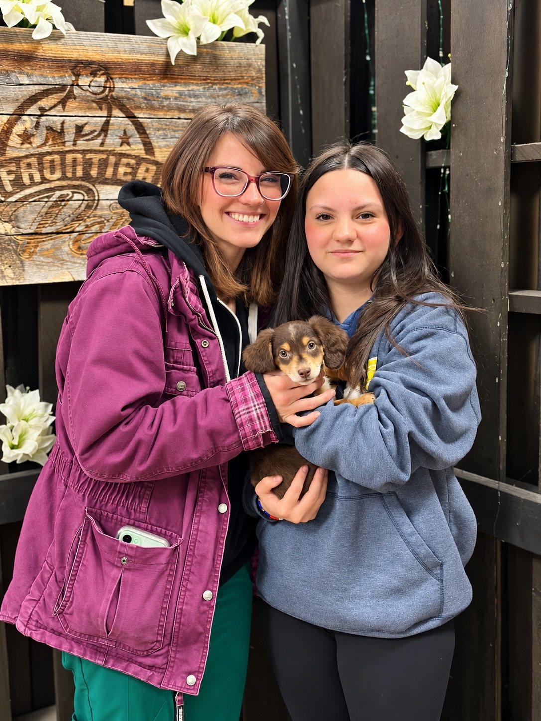 Two women smiling and holding a small brown and tan puppy in front of a Frontier Pets wooden sign
